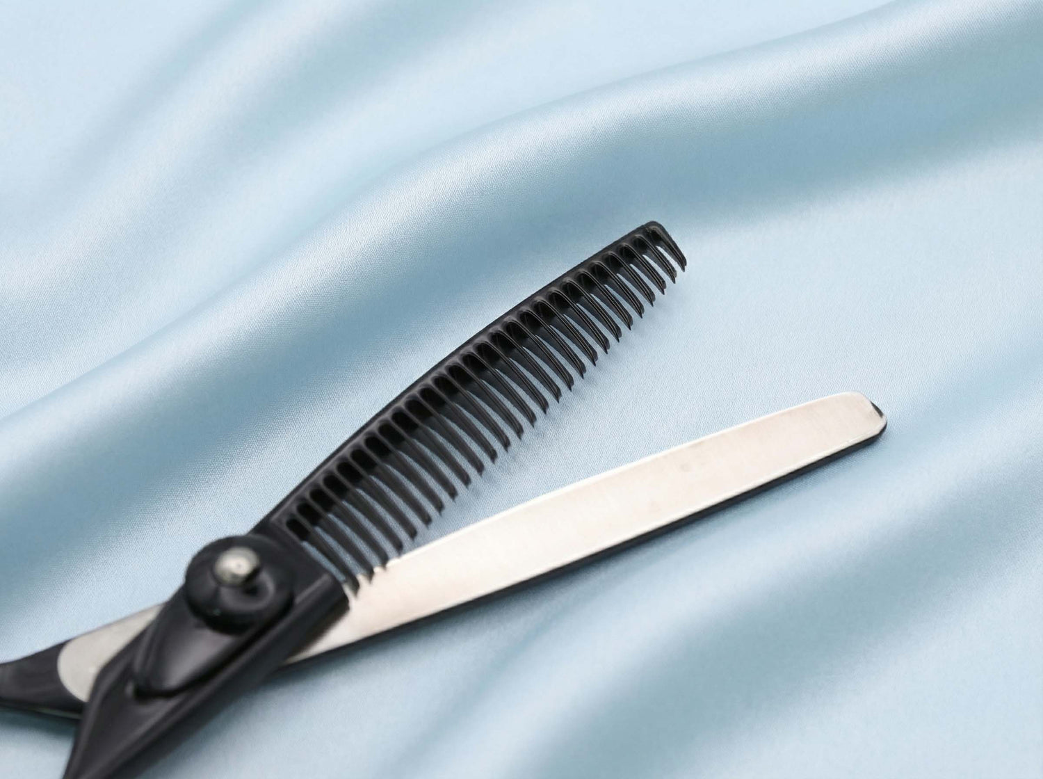 Schwarze Friseurschere mit Ausdünnungsklinge auf hellblauem, seidig schimmerndem Stoff liegend