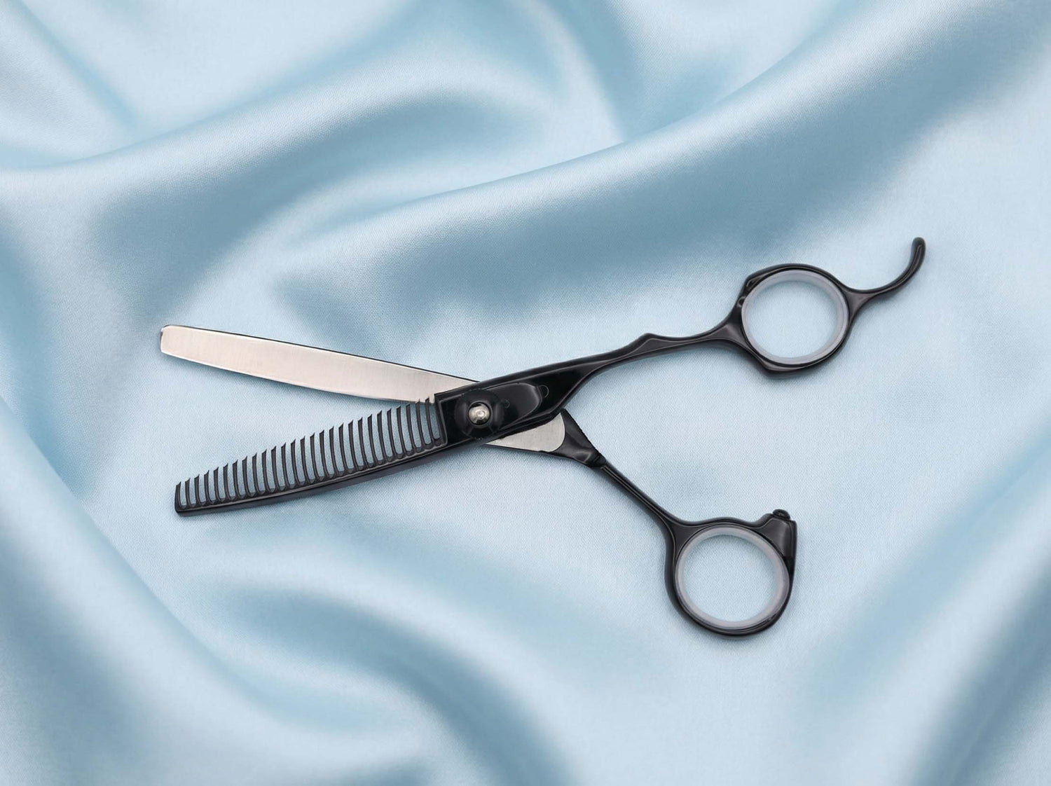 Schwarze Ausdünnungsschere mit geradem Griff und feiner Klinge auf hellblauem, glattem Stoff liegend