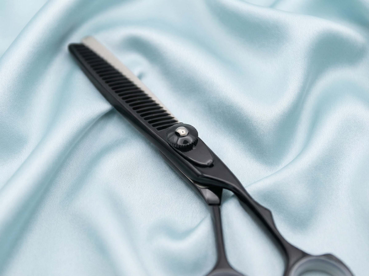 Schwarze Haarschneideschere mit Ausdünnungszähnen und verstellbarem Schraubverschluss auf hellblauem Stoff liegend