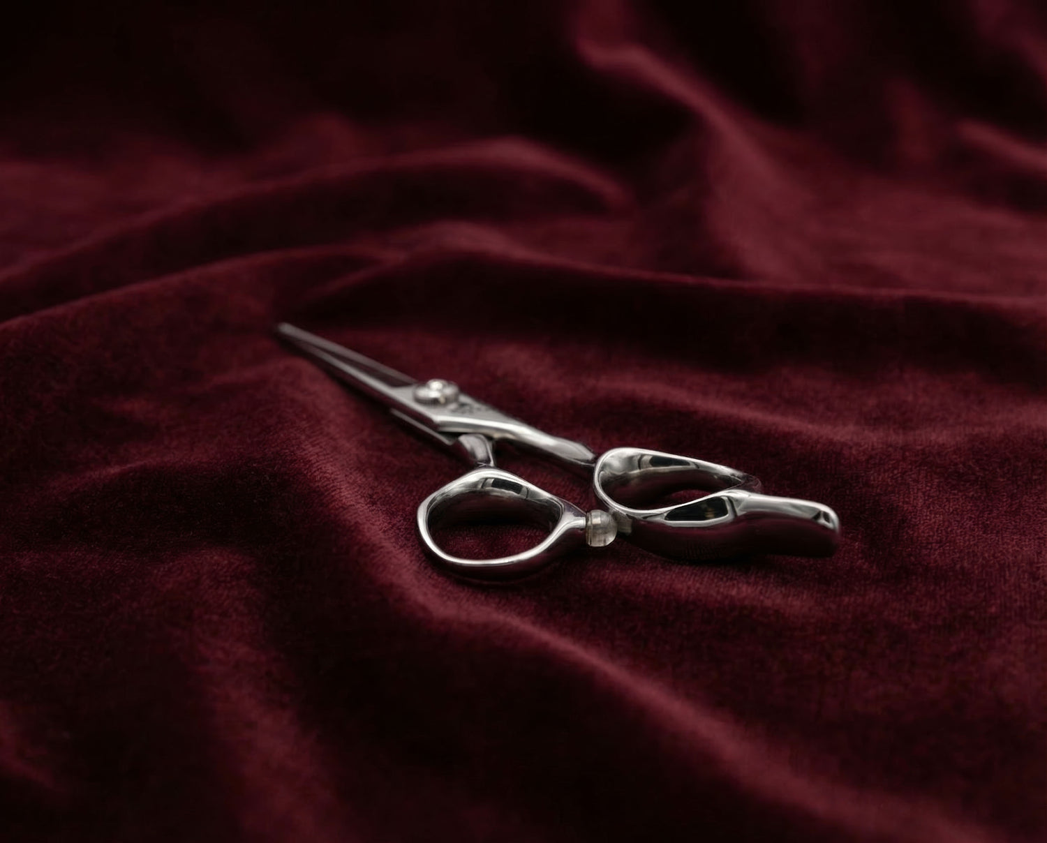 Friseurschere aus poliertem Edelstahl mit ergonomischem Griff liegt auf weichem, dunkelrotem Stoff