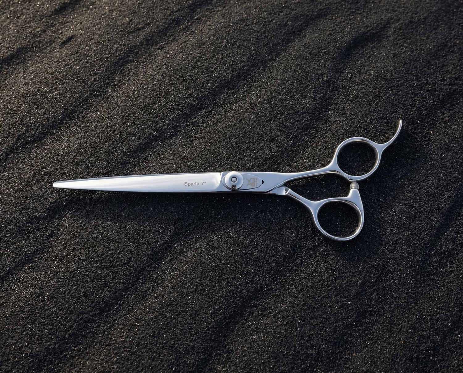 Glänzende Haarschneideschere mit ergonomischem Griff und filigraner Klinge liegt auf dunklem, körnigem Sandboden